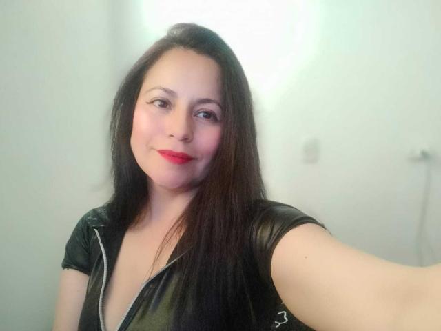 EkaterinaSex with long black hair, red lipstick, and a black leather shirt, smiling at the camera, extending her arm for a selfie. Her tits are partially visible.