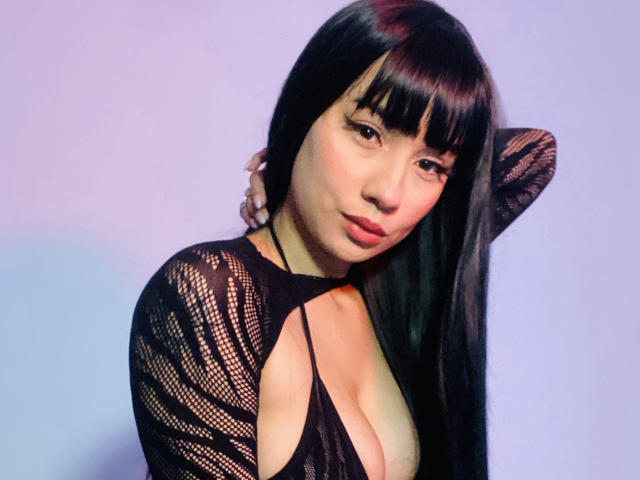 ivannity with long black hair, bangs, and a cute face, wearing a black mesh top that shows her tits, her arm behind her head, looking at the camera.