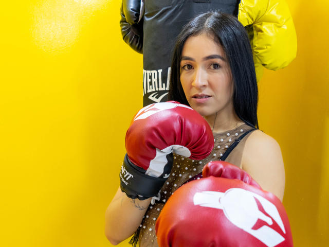 NicoColeman, Latina with long black hair, wearing a polka-dot dress and red boxing gloves, stands in front of a yellow wall with a yellow boxing glove and black bag.
