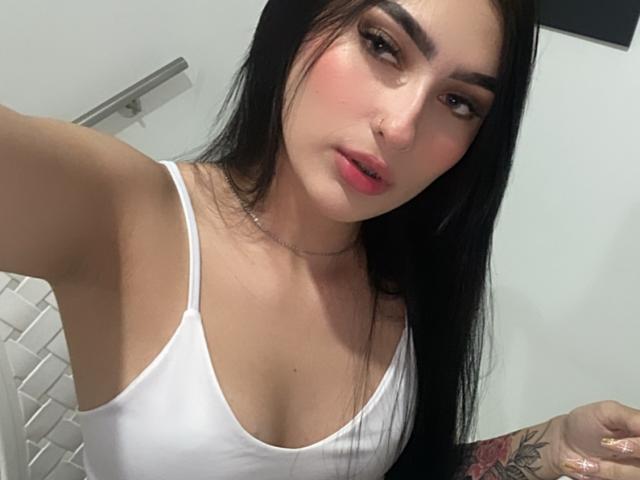 SevenJeyn met lang zwart haar, een wit tanktopje, roze lippen en een neusring, die een tatoeage op haar rechterarm laat zien, met een lichte glimlach en opgetrokken wenkbrauwen..