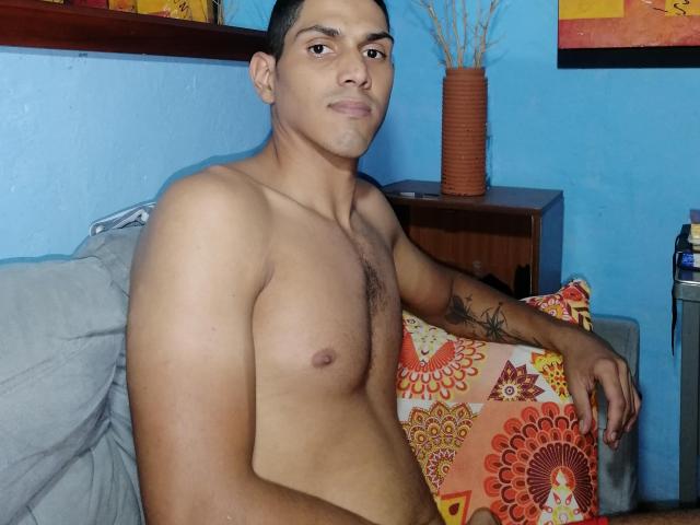 McBride, shirtless with a slight chest hair, tattoo on right arm, brown skin, short black hair, sitting on couch, arm on colorful pillow, blue wall, wood shelf in background.