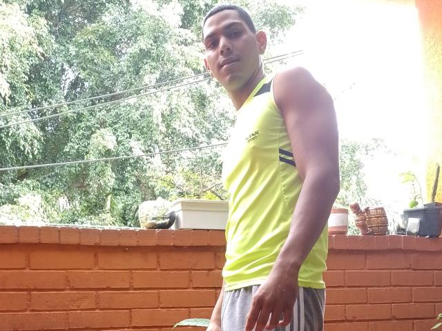McBride, a muscular dude with tan skin, short black hair, and a neon yellow sleeveless shirt, stands in front of a brick wall, wearing gray shorts.