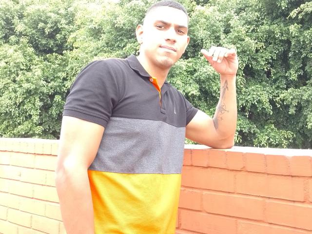McBride, a muscular dude with a tattoo on his forearm, wearing a black and yellow polo, leans on a brick wall, holding a cigarette with a smug expression.