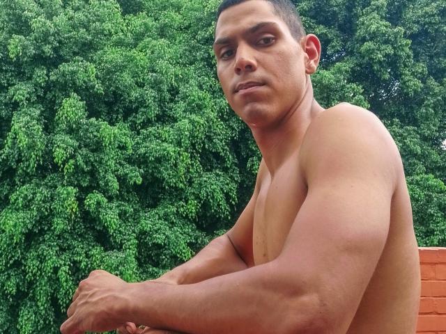 McBride's shirtless, muscular upper body with defined arms and a slight tan, green leafy background, short dark hair, small earring in left ear, looking to the side with a neutral expression.