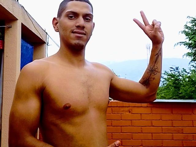 McBride, shirtless, has a toned, muscular build with small tits, a tattoo on his right arm, and short brown hair. He's making a peace sign, smiling, and has a goatee.