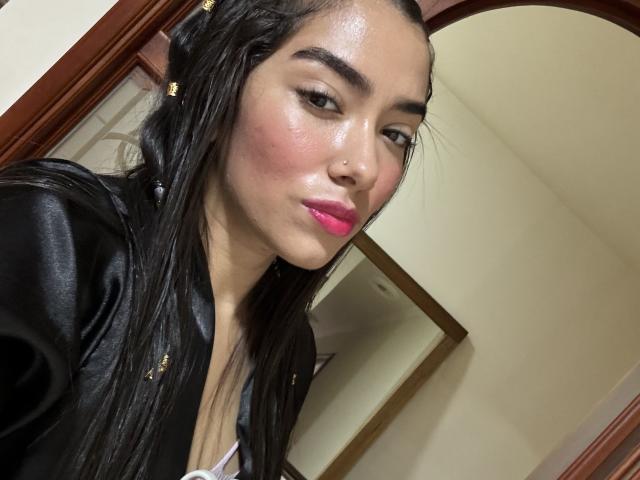 SharonKlart, con su cabello negro brillante, lápiz labial rosa y una chaqueta de cuero negro, mostrando su piel tersa y un pequeño piercing en la nariz, tomándose un selfie..