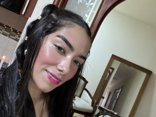 SharonKlart with shiny skin, pink lipstick, black hair in braids, smiling at camera, mirror reflection showing her face, brown wooden frame, white walls in background.
