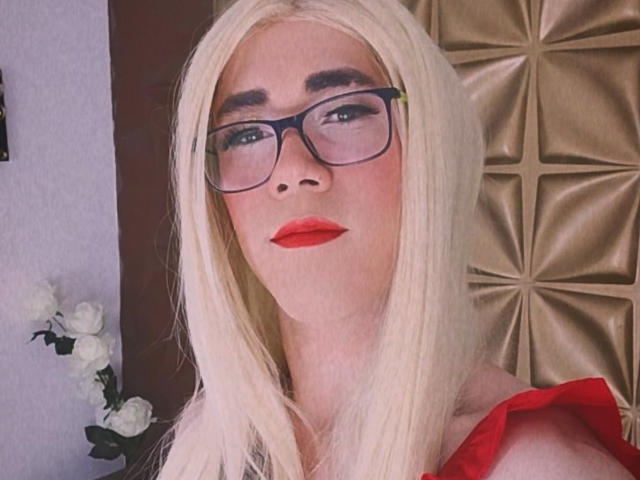 ElizabethQuenX with long blonde hair, red lipstick, and black glasses, looking slightly to the side, wearing a red top, has a cute face and is staring off confidently.