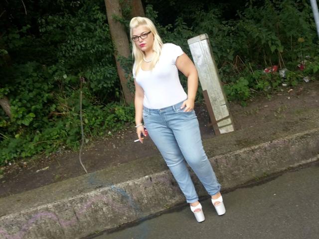 MariellaSun-hot with blonde hair, glasses, white tee, blue jeans, and white heels, holding a cigarette, standing on a sidewalk, hands in pockets, curvy with a medium ass.