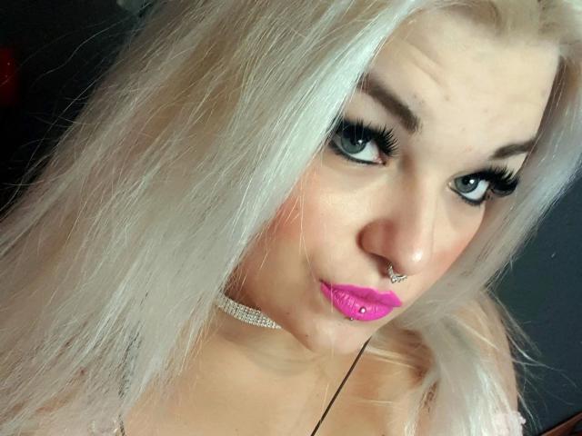MariellaSun-hot has platinum blonde hair, blue eyes, pink lipstick, a septum piercing, and a sparkly choker. Her tits are slightly visible. She's looking at the camera with a smirk.