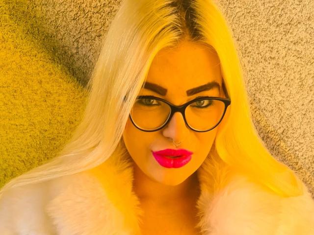 MariellaSun-hot with blonde hair, black glasses, pink lipstick, white fur coat, and a nose ring, looking directly at the camera with a slight smile.