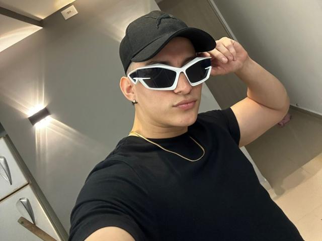 SantiagoXoxo with a black cap, white sunglasses, black t-shirt, gold chain, and a small ear piercing, looking down with a slight smirk. His ass is not visible.