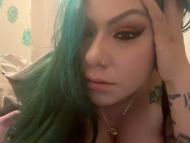 Blush69 with green-blue hair, thick eyebrows, winged eyeliner, nose ring, and tattoos, touching her head, wearing a necklace, and showing cleavage. Soft, serious expression.