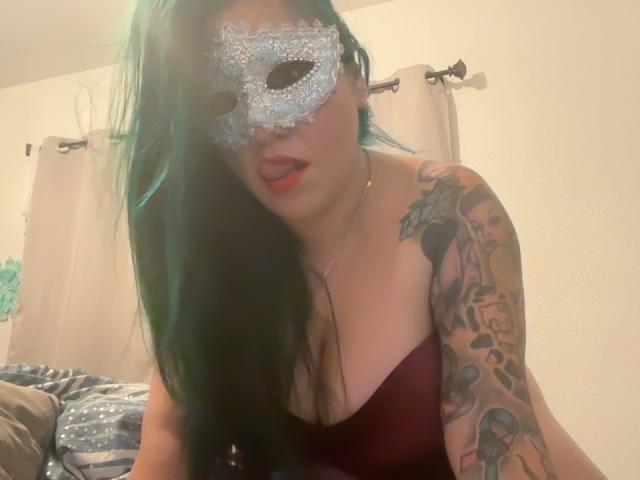 Blush69 with teal hair, wearing a glittery mask, red lipstick, and a deep cleavage in a black top, tattooed arm, leaning forward, bedroom setting, slightly blurred photo.