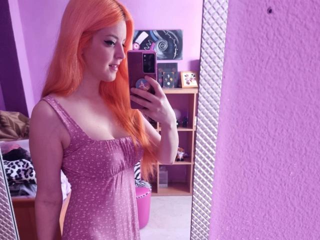 SaraKroft-hot with long orange hair, small tits, wearing a pink polka-dot dress, taking a mirror selfie in a purple room, smiling, holding a phone with a cute case.