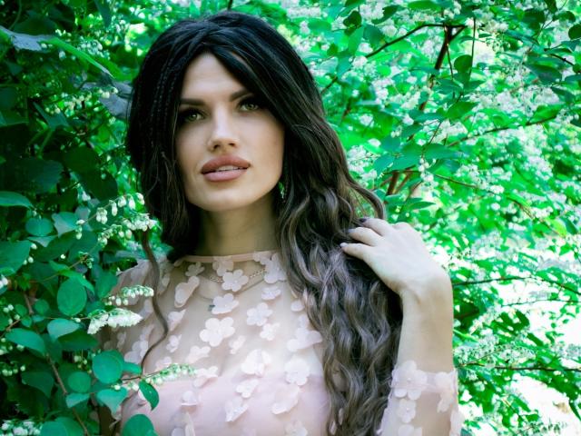 MilaLoona with wavy dark hair, wearing a sheer pink top with flower patterns, standing amidst green leafy branches, looking at the camera with a slight smile.