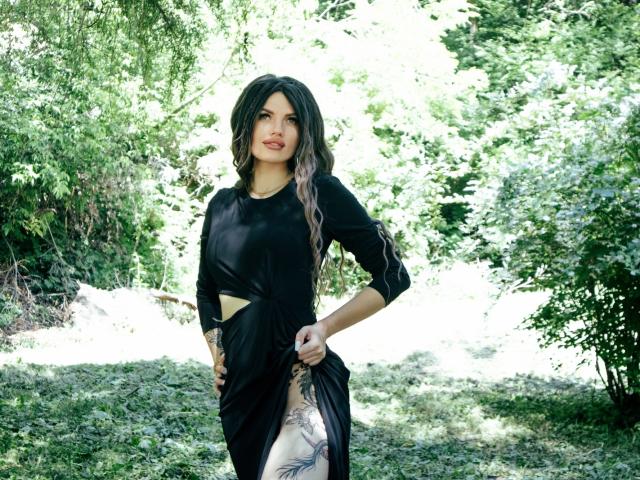MilaLoona with long black and purple hair, wearing a black dress, showing a tattoo on her thigh, standing in a sunny green forest, hands on dress, looking at camera.