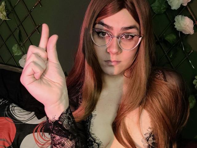 IsabelaCooper with long red hair, glasses, and lace top, showing a middle finger, has pale skin and small breasts, looking at the camera with a neutral expression.