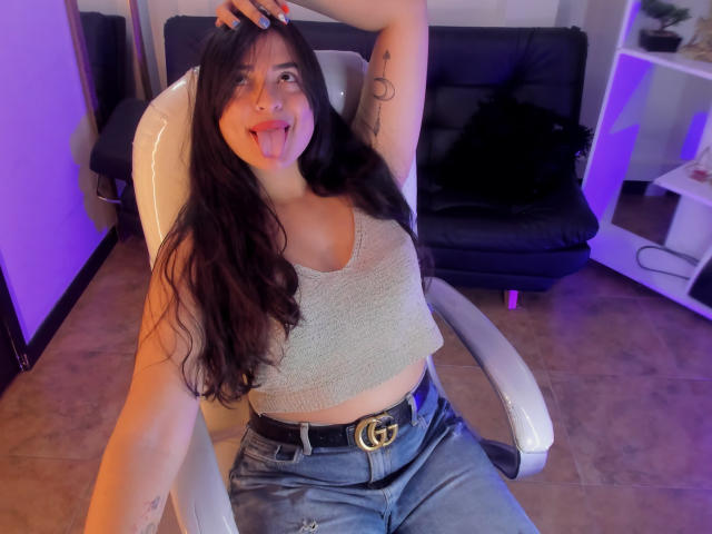 KylieFire with long dark hair, sticking out her tongue, wearing a beige crop top and blue jeans, sitting in a white chair, hand on head, visible arm tattoo.