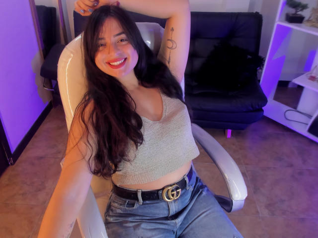 KylieFire with long black hair, smiling, wearing a beige crop top, blue jeans, and a Gucci belt, sitting in a white chair, arm raised, showing a small tattoo.
