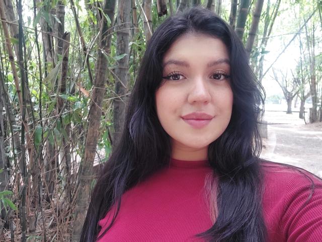 LauraRichis with long black hair, wearing a red ribbed shirt, has a curvy face, full lips, and brown eyes, standing in front of bamboo trees.