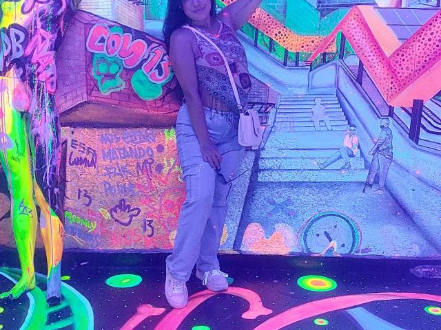 LauraRichis with medium tits, wearing a sparkly sleeveless top, light blue jeans, white sneakers, and a white crossbody bag, standing in a neon-lit, colorful urban graffiti setting.