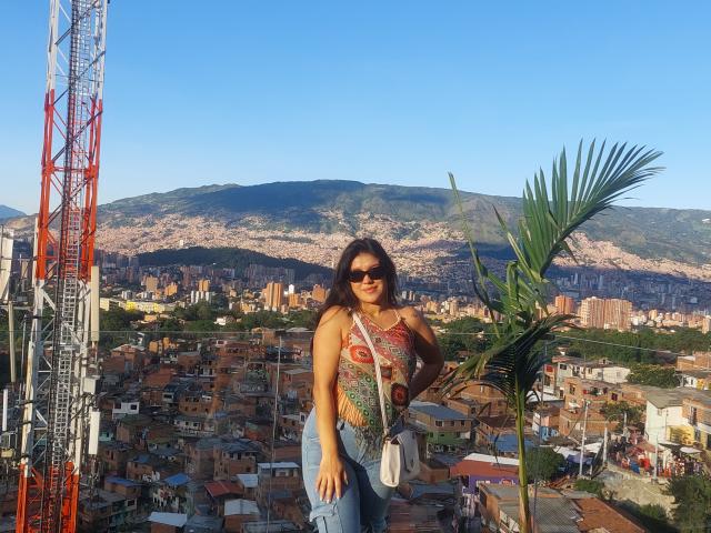 LauraRichis with tanned skin, wearing a colorful, patterned top, blue jeans, and sunglasses, standing in front of a tall red and white crane, with a palm tree to the right.