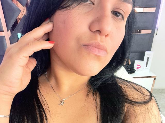 VivianaThompson has black hair, light skin, and is pouting with her lips slightly curled. She's touching her cheek with red-painted nails, wearing a silver necklace.