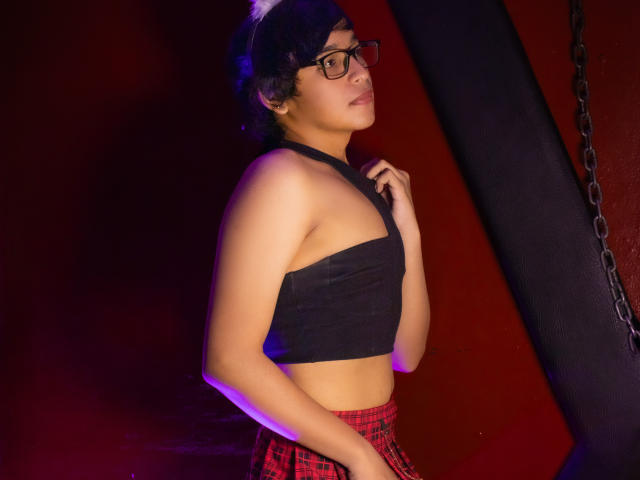 DannyHotz with short black hair, glasses, black tube top, red plaid skirt, standing, hand on chest, purple-blue lighting, looking right, red and black wall, chain in background.