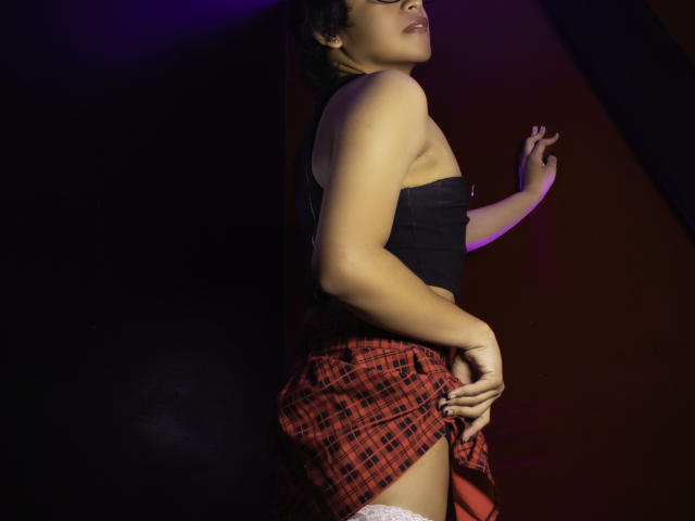 DannyHotz with short curly hair, black strapless top, red plaid skirt, white thigh-highs, hand on wall, ass slightly exposed, looking up, dark and purple-lit background.