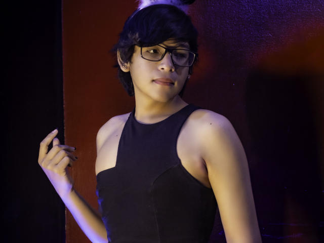 DannyHotz with short black hair, glasses, and black sleeveless top, holding a wall, looking slightly to the side, with a subtle smile, and a small tattoo on their hand.