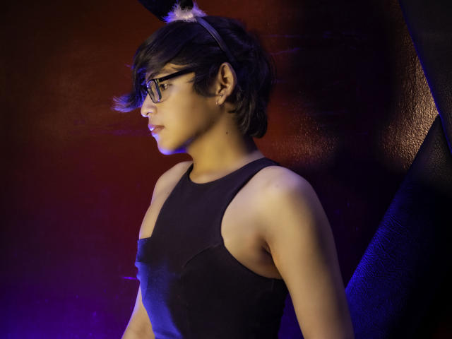 DannyHotz, wearing glasses and a black sleeveless top, has short black hair with a white puff, standing in profile with a serious expression, against a dark, textured background.