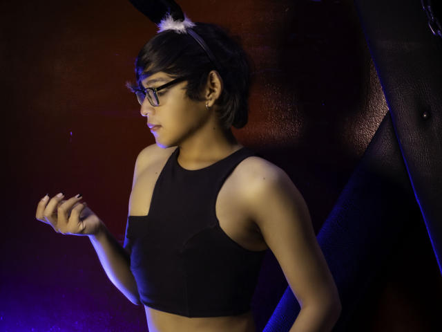 DannyHotz has short black hair, glasses, and a black sports bra, wearing a white headband with black ears, standing with a hand up, in a dark, blue-lit setting.