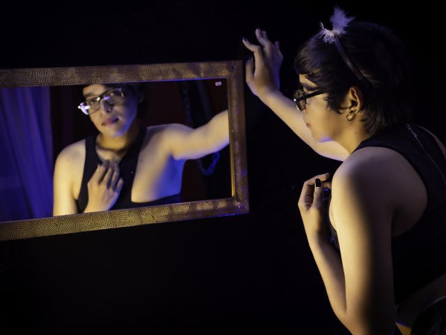 DannyHotz with short black hair, glasses, and a black top, staring at a mirror, touching it with one hand, and holding their chest with the other. Reflecting face.