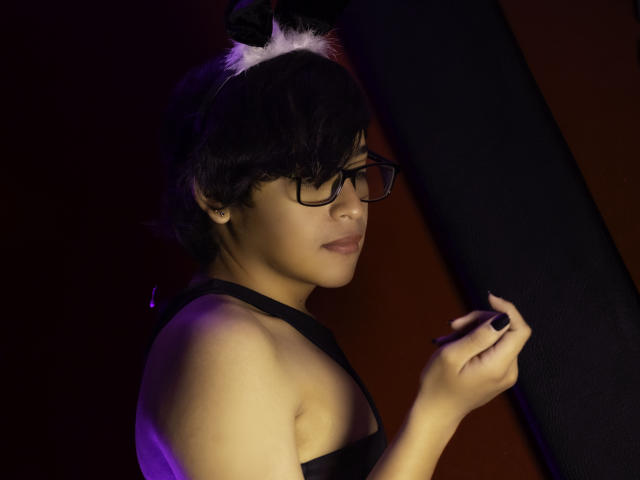 DannyHotz with short black hair, glasses, and white bunny ears, wearing a black strapless top, touches her nose, purple lighting, and dark background, showing a calm, sexy expression.