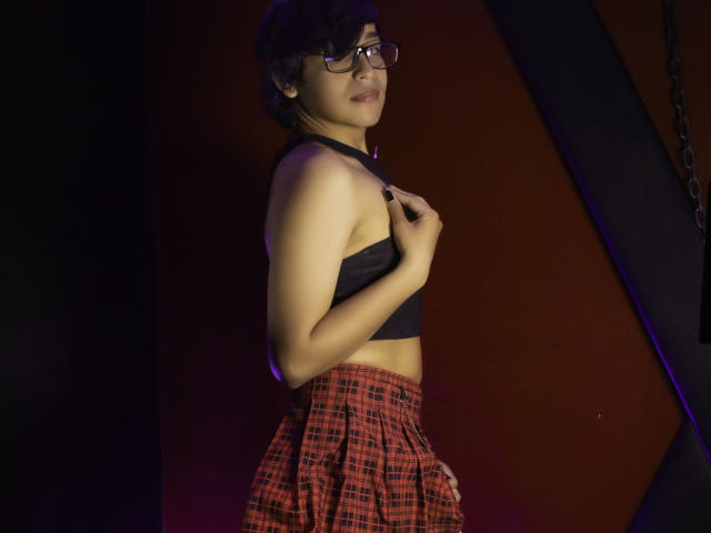DannyHotz with short black hair, glasses, black crop top, and red plaid skirt, standing, right side, hand on chest, confident, sexy, purple and red lighting.