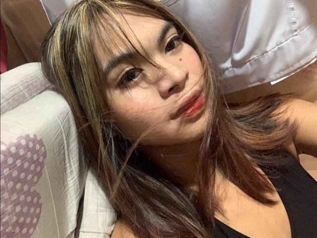 AsianBigBoobsBigCock with brown hair and blonde highlights, looking at camera, wearing black top, small red lipstick, white shirt in background, subtle smile, relaxed pose.