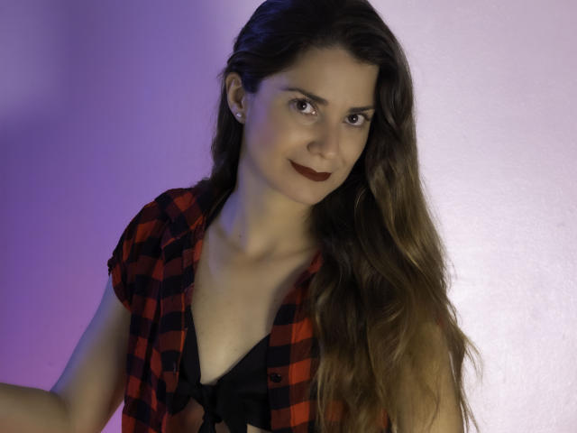 NatashaKute with long brown hair, red-black plaid shirt, black lace bra, and dark lipstick, smiling subtly, showing her tits slightly. Purple-blue lighting on her.
