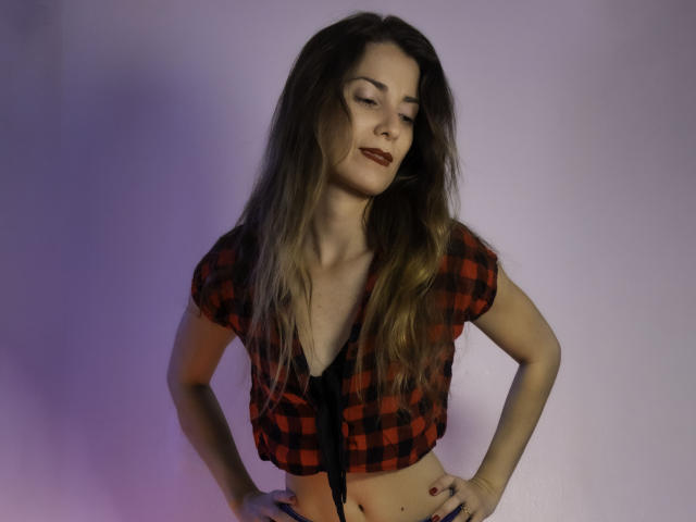 NatashaKute with long brown hair, red-black checkered crop top, hands on hips, slight smile, subtle makeup, exposed midriff, confident pose, purple-blue lighting.