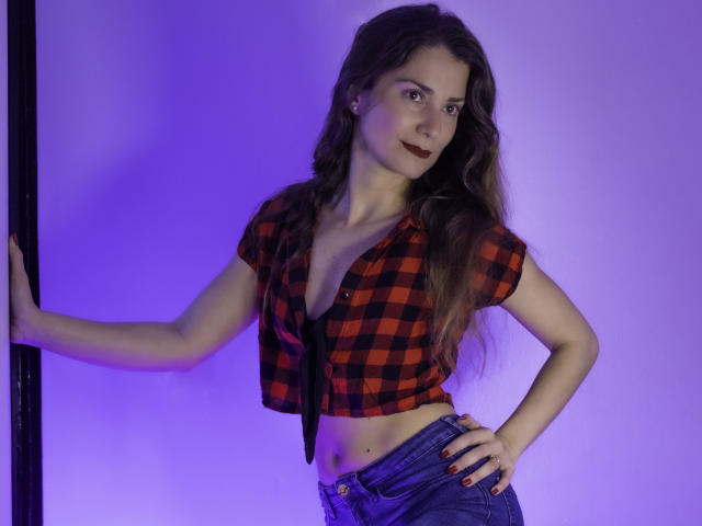 NatashaKute with long brown hair, wearing a red-black checkered crop top, exposing small tits, and blue jeans, standing with one hand on a pole, under purple lighting.