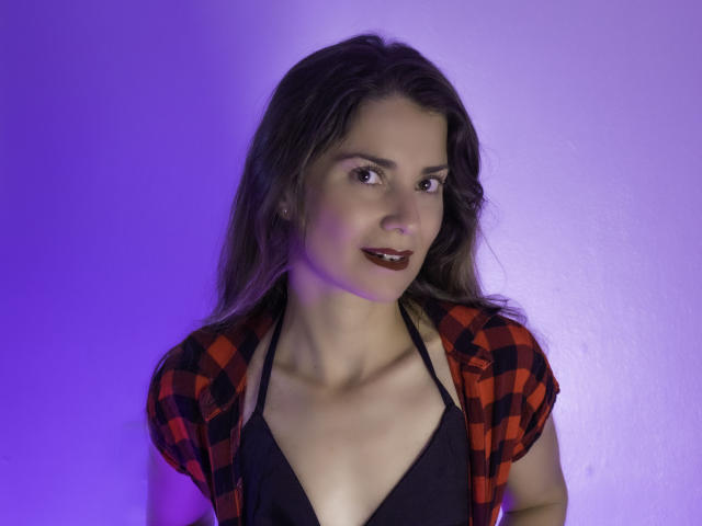 NatashaKute with long brown hair, red plaid shirt, black halter top, showing her tits, purple lighting, smiling with dark red lipstick.