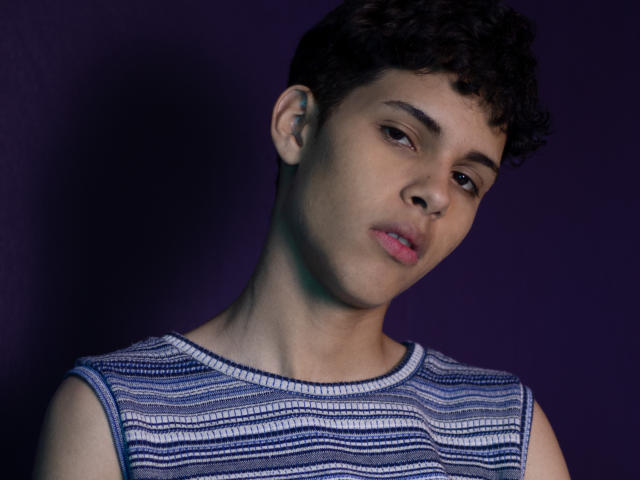 DenisBachh with curly dark hair, light brown skin, and a serious expression, wearing a blue and white striped sleeveless shirt, against a dark purple background.