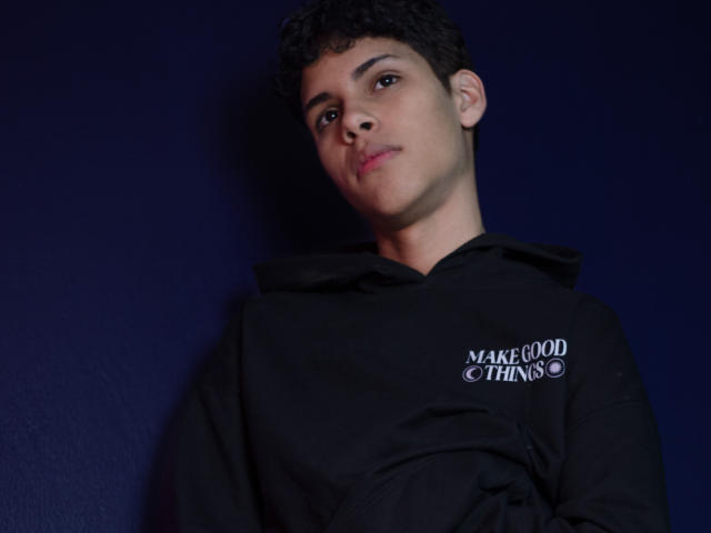 DenisBachh, short curly black hair, light brown skin, slightly cocky smile, wearing black hoodie, standing against dark blue wall, soft lighting, upper body shot.
