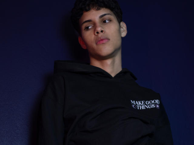DenisBachh with curly dark hair, light brown skin, and a serious expression, wearing a black hoodie with a white text print, standing against a dark blue background.