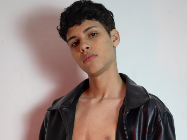 DenisBachh with curly black hair, brown eyes, and light brown skin, wearing a black leather jacket, looking slightly to the side with a neutral expression, no shirt underneath.