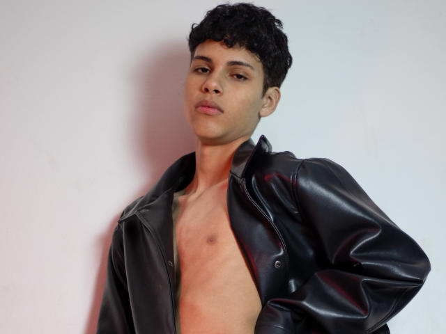 DenisBachh, with curly black hair, wearing a black leather jacket, is shirtless, showing a small bruise on his chest. He's leaning against a white wall, looking serious.