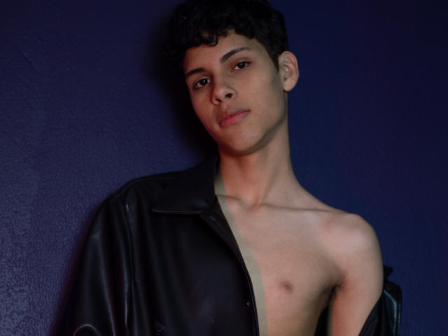 DenisBachh with curly black hair, light brown skin, and a slight smile, wearing an open black leather jacket, exposing a small, flat chest against a dark blue wall.
