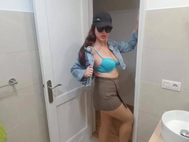 SensualClaudine-hot stands in a door, wearing a black cap, sunglasses, denim jacket, blue lace bra, and brown skirt, showing her tits and a slightly open door.