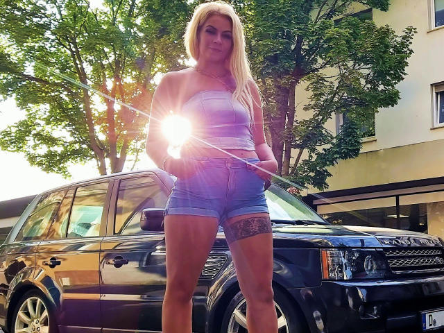 GeilesBabe-hot with long blonde hair, striped tank top, and denim shorts, holding a sun flare, standing in front of a black SUV with a visible thigh tattoo.