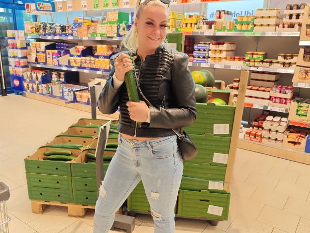 GeilesBabe-hot with blonde hair, wearing a black leather jacket, ripped blue jeans, holding a green cucumber, standing in a grocery store with stacked green boxes.